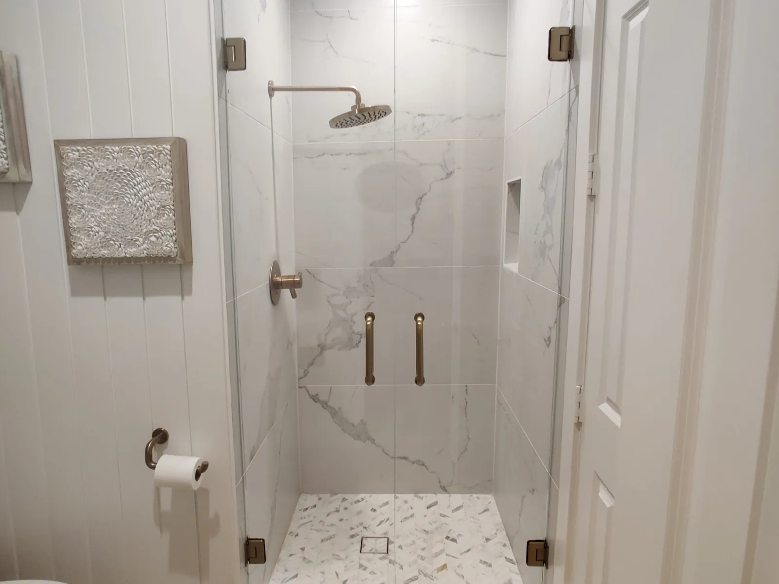 Pair of frameless glass shower doors with brushed bronze hinges and pulls in a white marble-look shower