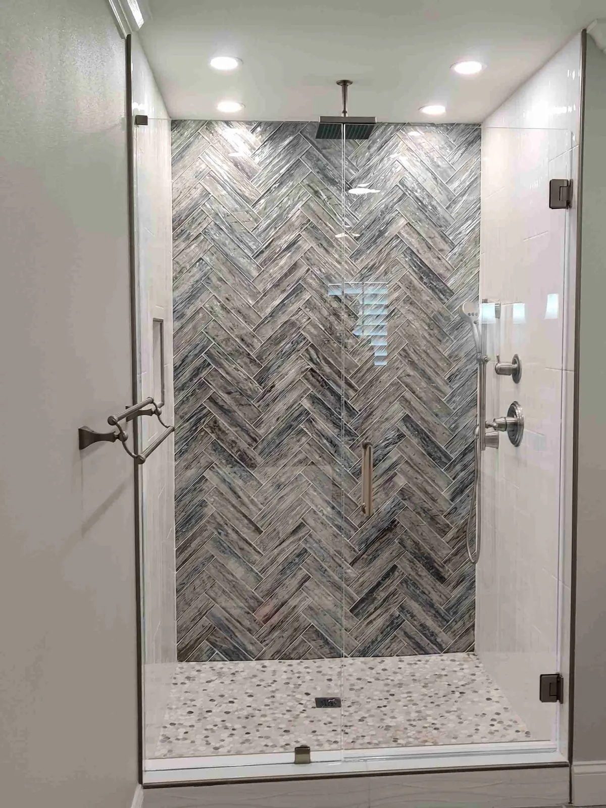 Frameless glass shower door and fixed panel in front of a blue-gray herringbone accent wall and pebble floor