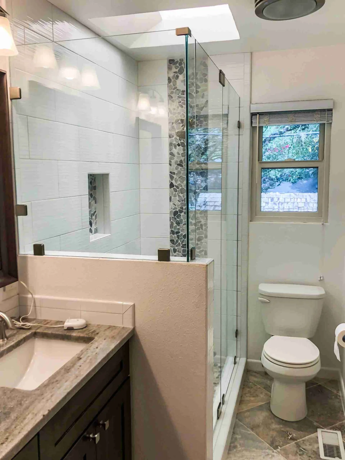 Frameless glass door and panel on a tiled knee wall beside a vanity, with a pebble tile accent strip