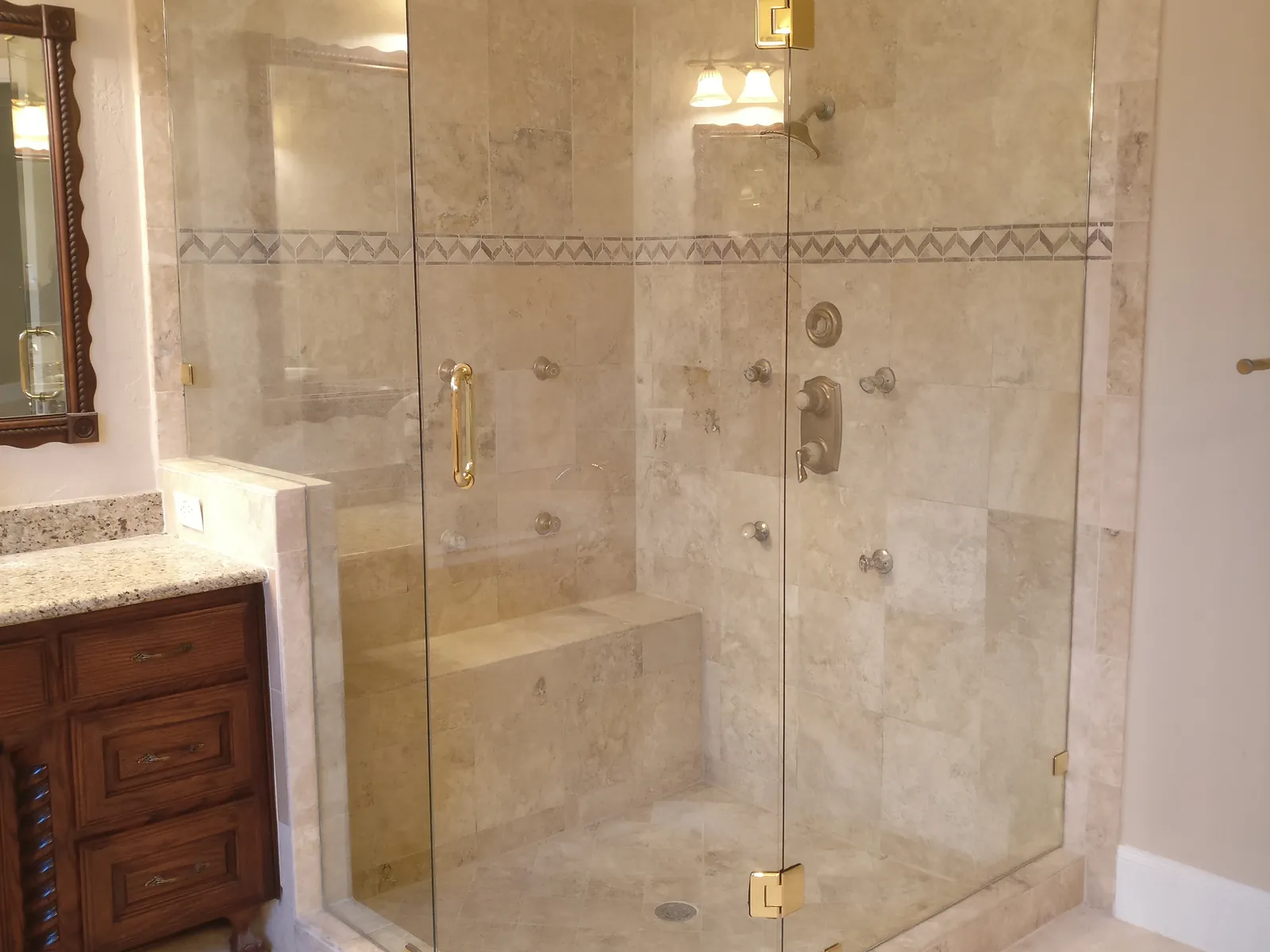 Neo-angle frameless glass shower with brass hardware, travertine-look tile and a built-in bench